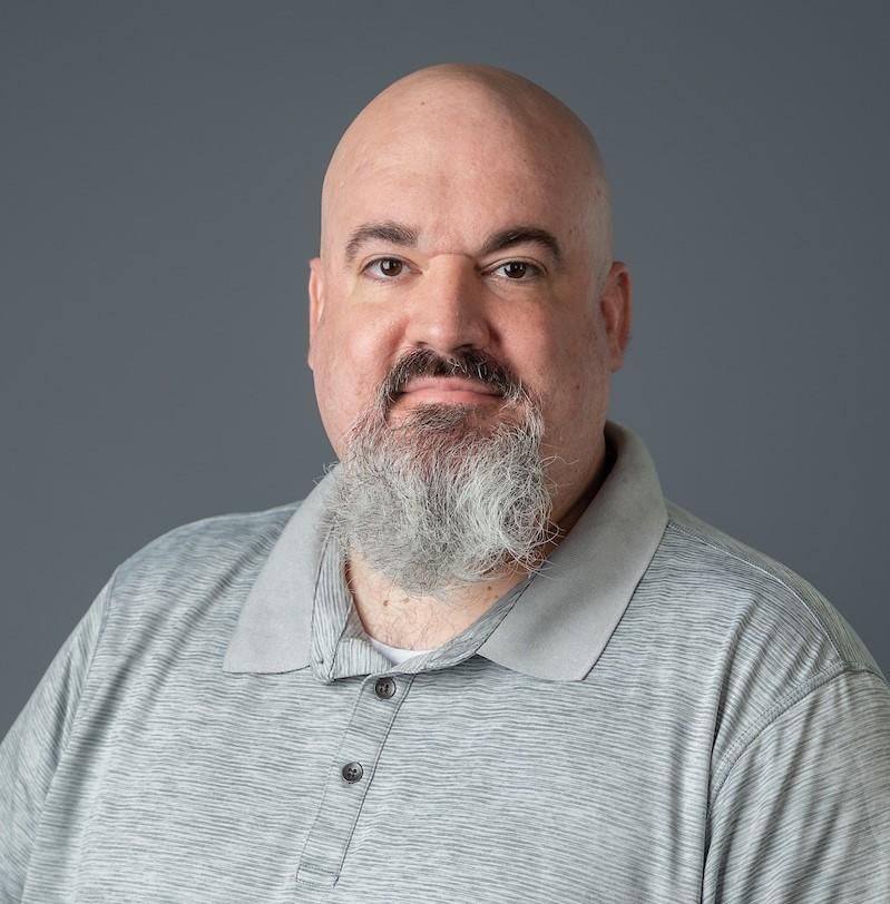 A man with a gray beard wearing a grey polo shirt poses for the camera.