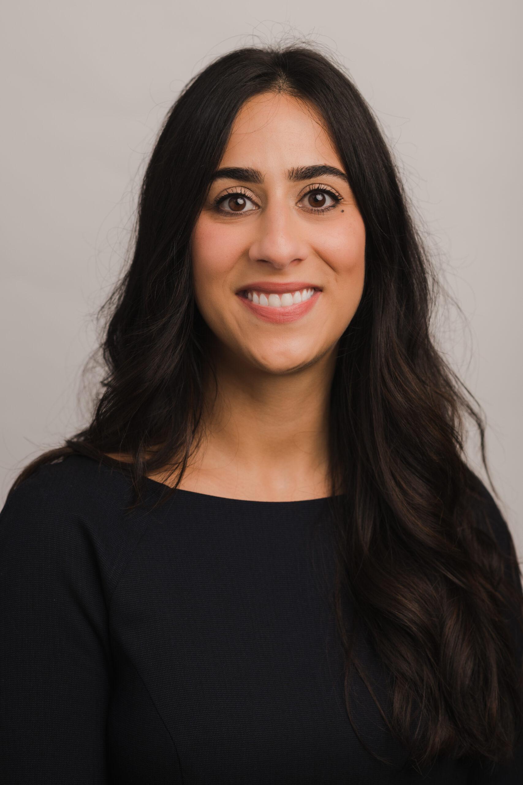 Layla lee stands in front of a gray background. she is smiling at the camera and is wearing a black top.