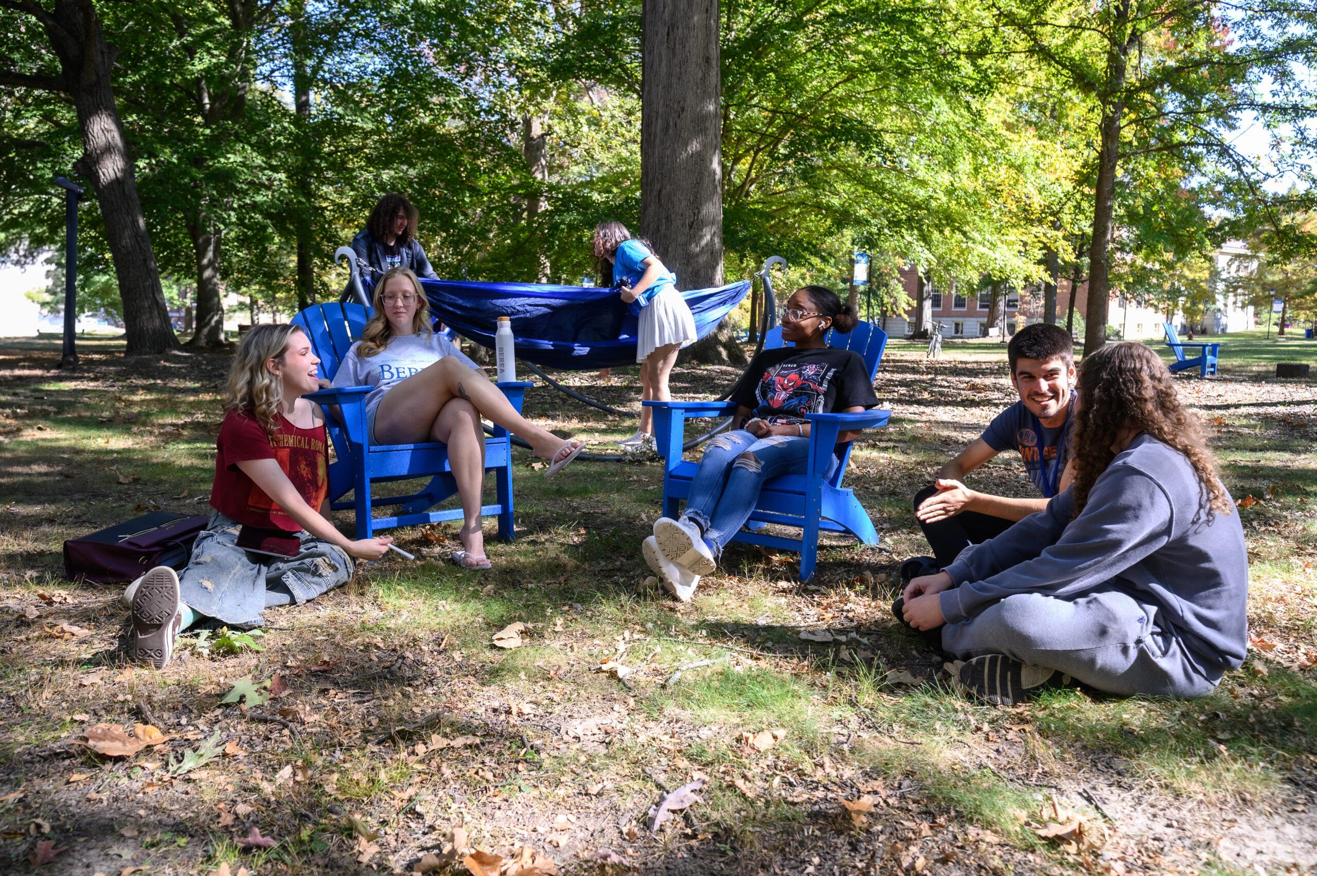 Five students are shown sitting on the quad in blue Adirondack chairs and in the grass, talking and laughing with one another.